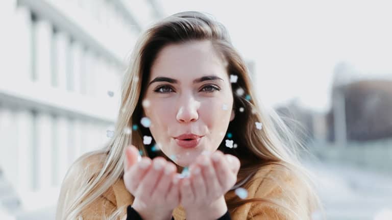 Eine Frau mit langen blonden Haaren und einem hellbraunen Pullover pustet glitzernde Konfetti aus ihren Händen in Richtung Kamera und verkörpert damit Freude. Für einen frischen Atem in solch heiteren Momenten entdecken Sie hilfreiche Tipps gegen Mundgeruch, um im Freien selbstbewusst zu bleiben.