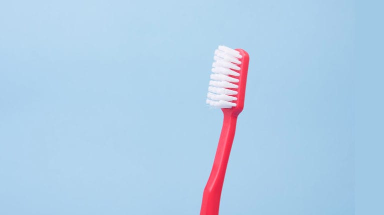 Eine rote Zahnbürste mit weißen Borsten steht aufrecht vor einem hellblauen Hintergrund. Die manuelle Zahnbürste ist mittig platziert und isoliert, ohne weitere Gegenstände im Bild.