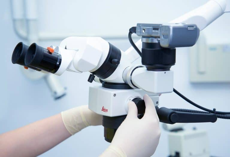 Eine Person mit weißen Latexhandschuhen stellt die Einstellräder eines weißen wissenschaftlichen Mikroskops ein, das in der Endodontie verwendet wird, mit binokularen Okularen und angeschlossener Kameraausrüstung, die in einer sauberen, hell erleuchteten Laborumgebung aufgestellt ist.