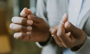 Eine Nahaufnahme von zwei Händen, die Zahnseide halten, wobei die Zahnseide fest zwischen den Fingern gespannt ist. Dieses tägliche Ritual ist ideal für die Prophylaxe und hilft, die Zähne zu pflegen und unterstützt ein effektives Biofilmmanagement. Der graue Oberteil und der unscharfe Hintergrund lenken den Fokus auf die Handlung.