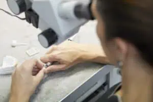 Eine Person benutzt ein Mikroskop, während sie an einem Labortisch mit zahnmedizinischen Werkzeugen arbeitet. Auf der grauen Oberfläche sind zahnmedizinische Formen und kleine Instrumente zu sehen, die auf eine präzise zahnmedizinische oder kieferorthopädische Arbeit hinweisen, die gerade durchgeführt wird. Das Gesicht der Person ist nicht deutlich zu erkennen.
