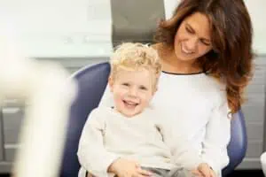 Eine lächelnde Frau mit gewelltem braunem Haar sitzt in einem Zahnarztstuhl und hält einen fröhlichen Jungen auf ihrem Schoß. Beide wirken glücklich und entspannt und spiegeln die fürsorgliche Atmosphäre der Kinderzahnheilkunde in einer hellen, sauberen Praxisumgebung wider.