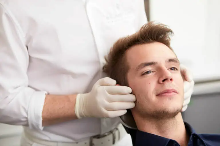 Ein junger Mann lehnt sich entspannt in einem Stuhl zurück, während eine Person mit weißen Handschuhen und weißem Kittel sanft seinen Kiefer und Nacken hält, was auf eine medizinische oder chiropraktische Untersuchung in einem klinischen Umfeld hindeutet.