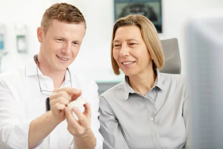 Ein Mann und eine Frau in Berufskleidung sitzen lächelnd zusammen. Der Mann hält ein Zahnmodell und erklärt der Frau die Implantologie. Beide wirken beschäftigt in dem hellen, modernen Büro des Zahnzentrums Kronberg im Taunus.