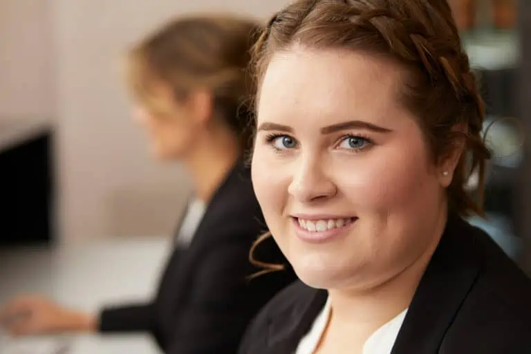 Eine junge Frau mit geflochtenem Haar und einem schwarzen Blazer lächelt in die Kamera. Sie hat helle Haut und blaue Augen. Im unscharfen Hintergrund sitzt eine andere Frau in ähnlicher Geschäftskleidung und konzentriert sich auf einen Computer. Die Umgebung scheint ein Büro zu sein.