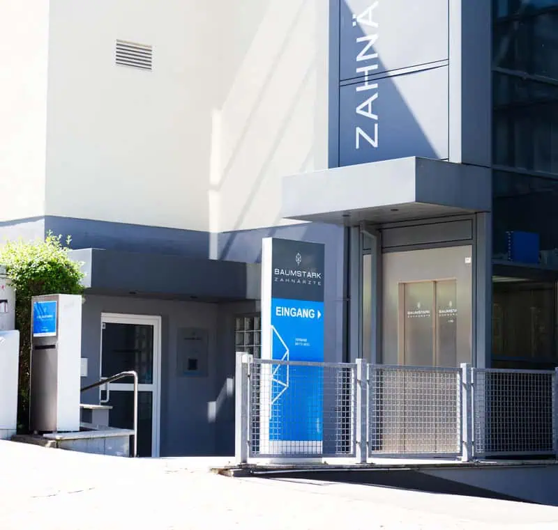 Ein moderner Zahnarzteingang zur Praxis Baumstark mit Glastüren und Schildern. Auf dem Gebäude steht "ZAHNÄRZTE", und ein blaues Schild "BAUMSTARK ZAHNÄRZTE EINGANG" begrüßt Neupatienten bei Zahnarzt Kronberg mit Fahrstuhl und ein Geländer, das zur Tür führt.
