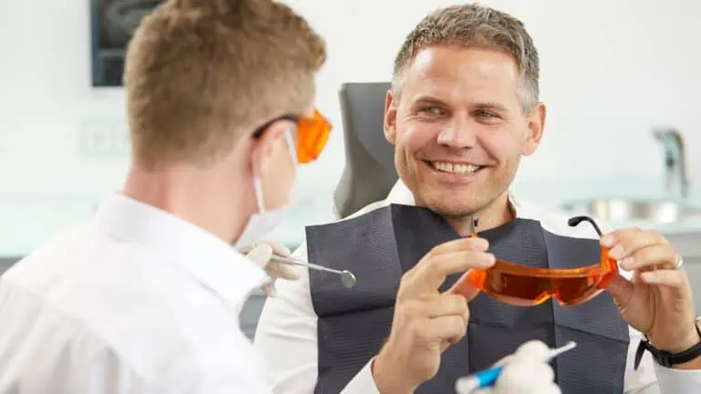 Ein Mann sitzt in einem Zahnarztstuhl, lächelt und trägt eine orangefarbene Schutzbrille. Ein Zahnarzt, der ebenfalls eine orangefarbene Brille und einen weißen Kittel trägt, spricht mit ihm, während er zahnärztliche Werkzeuge in der Hand hält. Beide wirken entspannt in einer hellen, modernen Zahnarztpraxis.
