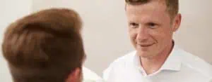 Ein Mann in einem weißen Hemd, mit einem warmen zahnbalance Lächeln, schaut eine andere Person an, die meist unscharf und von hinten gesehen ist. Die Atmosphäre wirkt freundlich, mit einem neutralen, hellen Hintergrund.