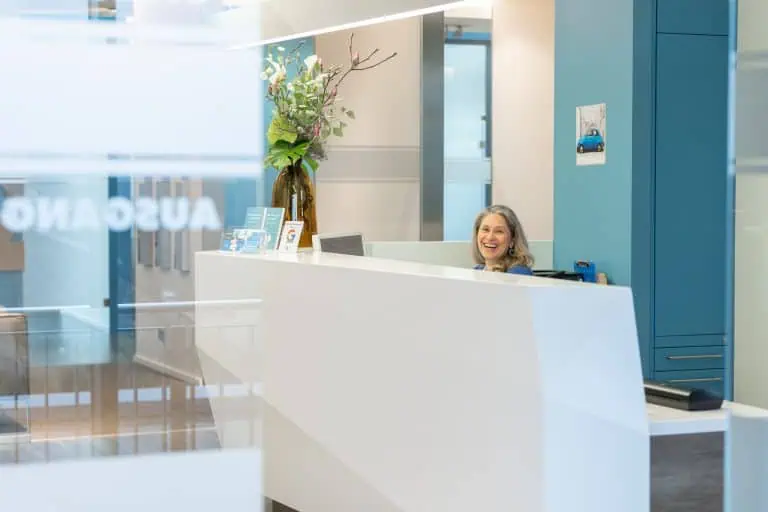 Eine Frau mit grauem Haar lächelt, während sie hinter einem modernen weißen Empfangsschreibtisch in einer Bürohalle sitzt. Auf dem Tresen stehen eine hohe Vase mit Blumen und Broschüren, die eine einladende, saubere Atmosphäre schaffen, die perfekt für jede Empfangsstellenanzeige ist.