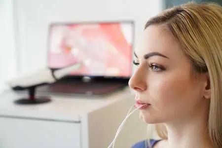 Eine Frau mit blondem Haar sitzt in einer DMD Untersuchung. Im Hintergrund ist ein unscharfer Computermonitor zu sehen, der ein medizinisches Bild in Großaufnahme zeigt.