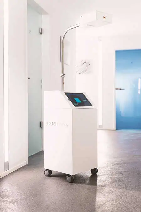Ein hohes, weißes, kastenförmiges medizinisches Gerät für digitale Kiefergelenkmessung auf Rädern mit einer Touchscreen-Oberfläche und einem erhobenen Arm, der in einer Leuchte oder einem Sensor endet, steht in einem modernen, hellen Flur mit hellen Wänden, Glastüren und einer blauen Tür im Hintergrund.