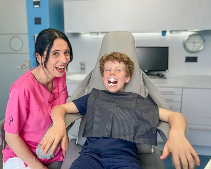 Ein lächelnder Zahnarzt in rosa Kitteln steht neben einem Jungen, der auf einem Zahnarztstuhl sitzt. Die spielerische Szene in einer hellen, modernen Klinik mit sichtbaren Geräten unterstreicht die sanfte Herangehensweise der Kinderprophylaxe Kronberg an die zahnmedizinische Versorgung von Kindern.