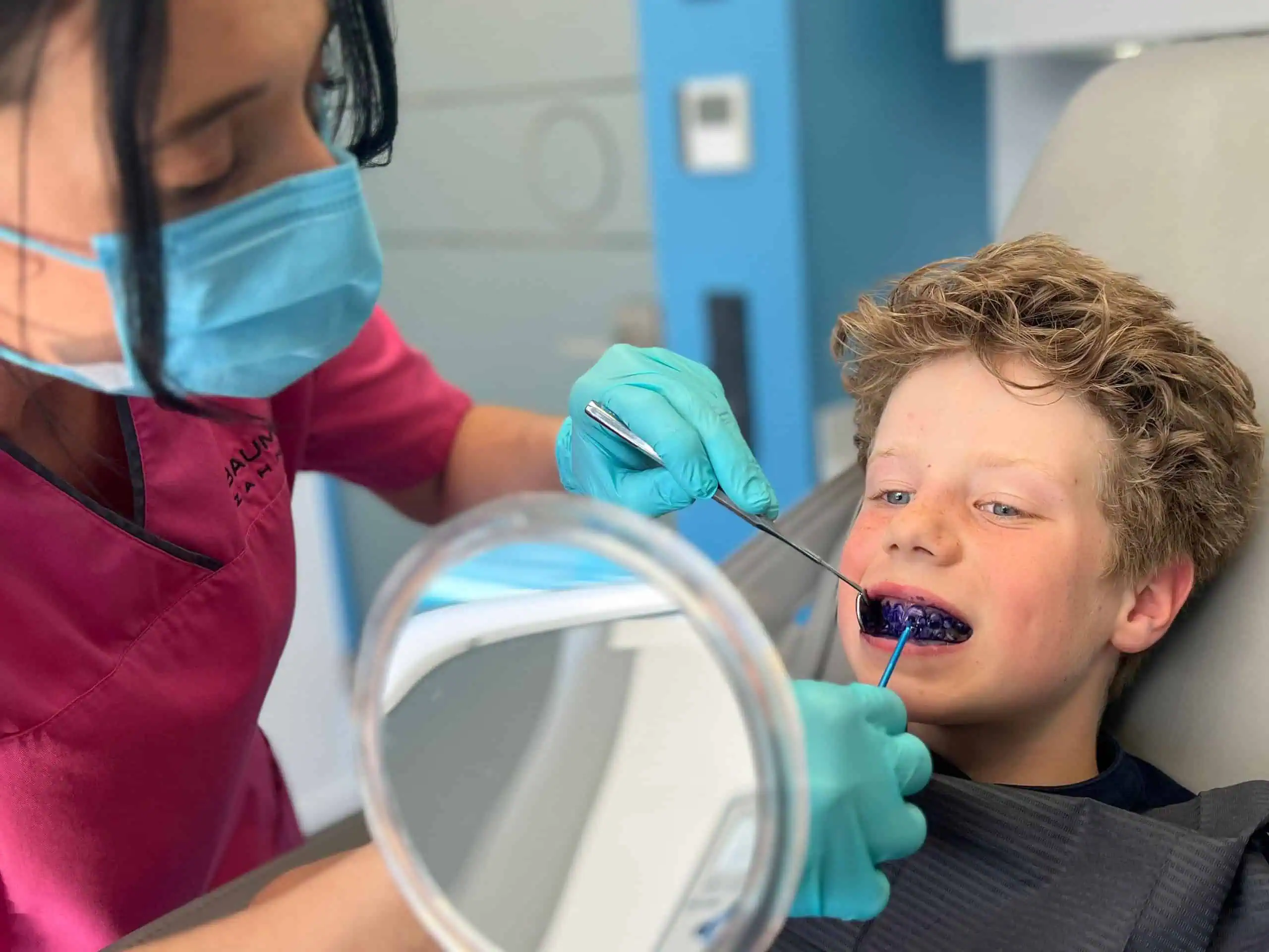 Eine Zahnärztin in rosa Kittel und blauer Maske behandelt die Zähne eines lächelnden Jungen mit einer blauen Zahnbehandlung. Der Junge, der im Rahmen der Kinderprophylaxe Kronberg zu Besuch ist, sitzt auf dem Zahnarztstuhl, hält einen Spiegel in der Hand und beobachtet, wie die Zahnärztin mit blauen Handschuhen arbeitet.