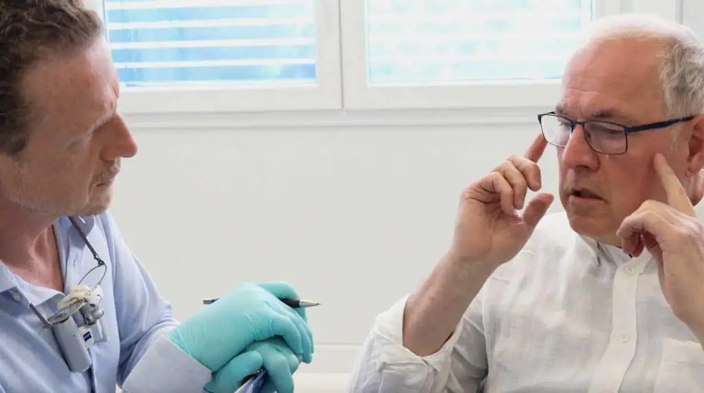 Zahnarzt Dr. Baumstark mit blauen Handschuhen und einer Zahnarztlupe bespricht mögliche CMD-Symptome mit einem älteren Mann mit Brille und weißem Hemd, der sich besorgt an die Schläfen fasst, während die beiden drinnen in der Nähe eines Fensters sitzen.
