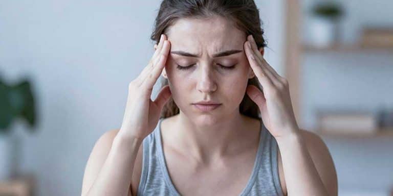 Eine junge Frau mit braunen Haaren, die ein graues Trägertop trägt, sitzt drinnen, die Augen geschlossen und die Augenbrauen zusammengezogen, und drückt ihre Finger gegen die Schläfen, als würde sie mit Kopfschmerzen oder Verspannungen im Kiefer zu kämpfen haben. Der Hintergrund ist leicht unscharf.