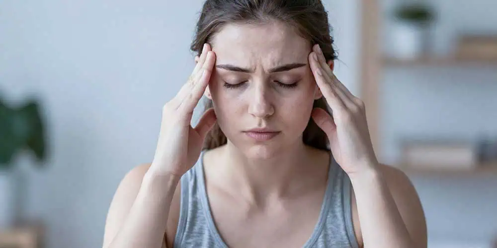 Eine junge Frau mit braunen Haaren, die ein graues Trägertop trägt, sitzt drinnen, die Augen geschlossen und die Augenbrauen zusammengezogen, und drückt ihre Finger gegen die Schläfen, als würde sie mit Kopfschmerzen oder Verspannungen im Kiefer zu kämpfen haben. Der Hintergrund ist leicht unscharf.