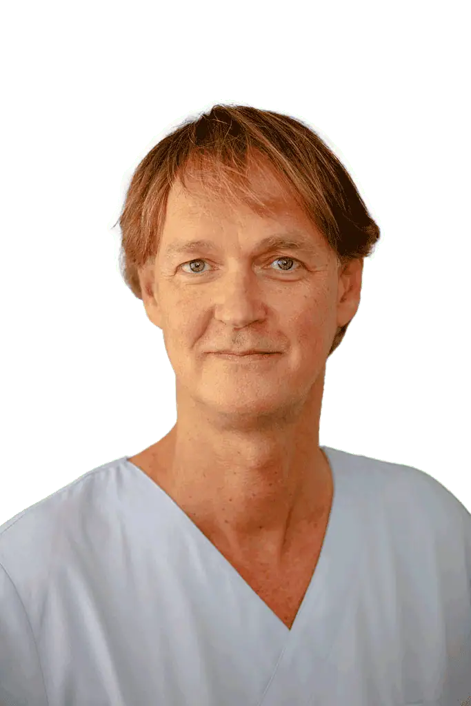 Eine Person mittleren Alters mit heller Haut, kurzen braunen Haaren und blauen Augen trägt ein hellblaues OP-Oberteil. Sie blickt nach vorne und lächelt leicht vor einem schlichten, hellen Hintergrund und vermittelt so den professionellen Eindruck eines Oralchirurgen aus Kronberg.